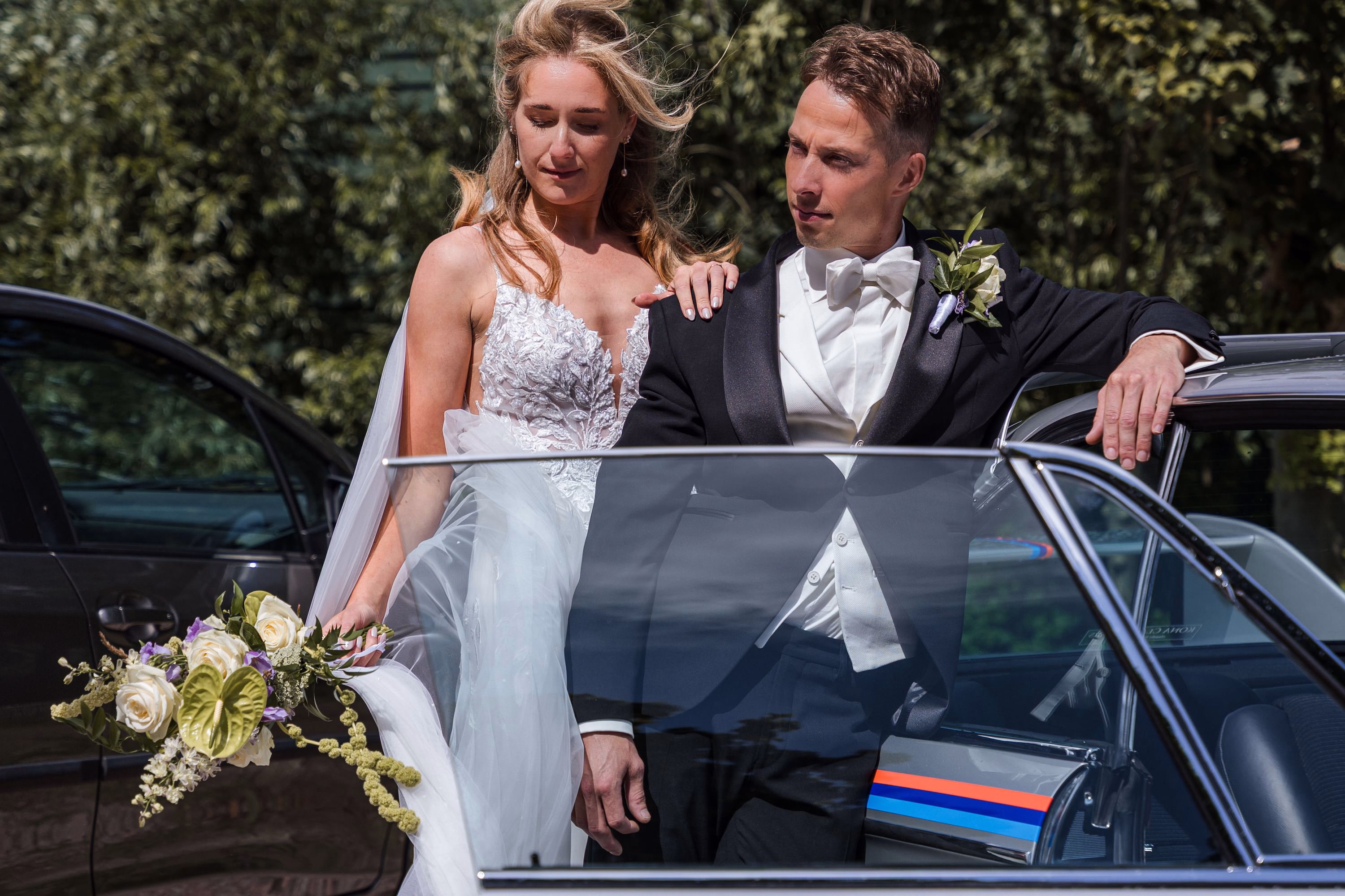 Trouwfotografie Zuid-Holland — bruidspaar poserend voor trouwauto met boeket